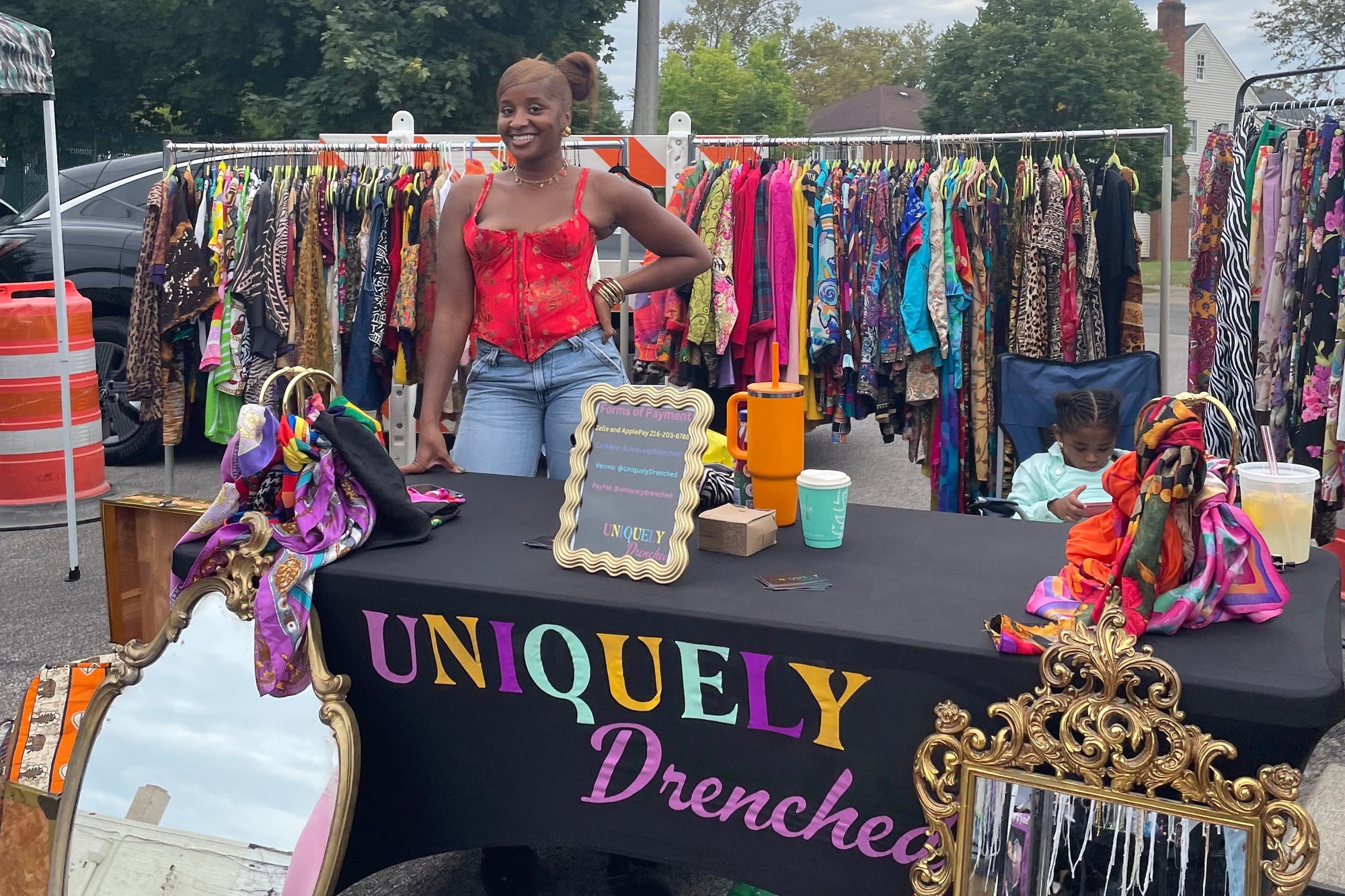 Person sitting at a street vendor stall with various items on display, including clothing and decorative mirrors.